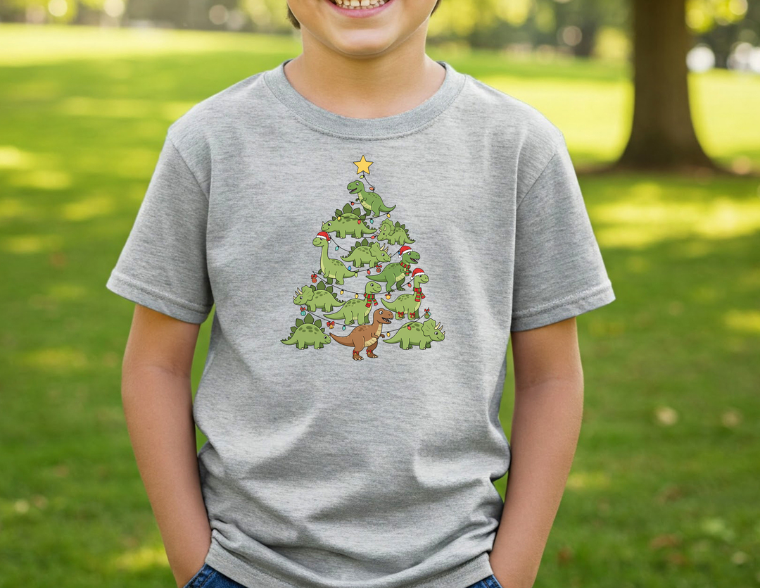 Child wearing Dino Tree Kids Tee with cartoon dinosaur Christmas tree graphic, hands in jeans pockets, smiling on a sunny day. Heather-gray, durable, and perfect for everyday use.