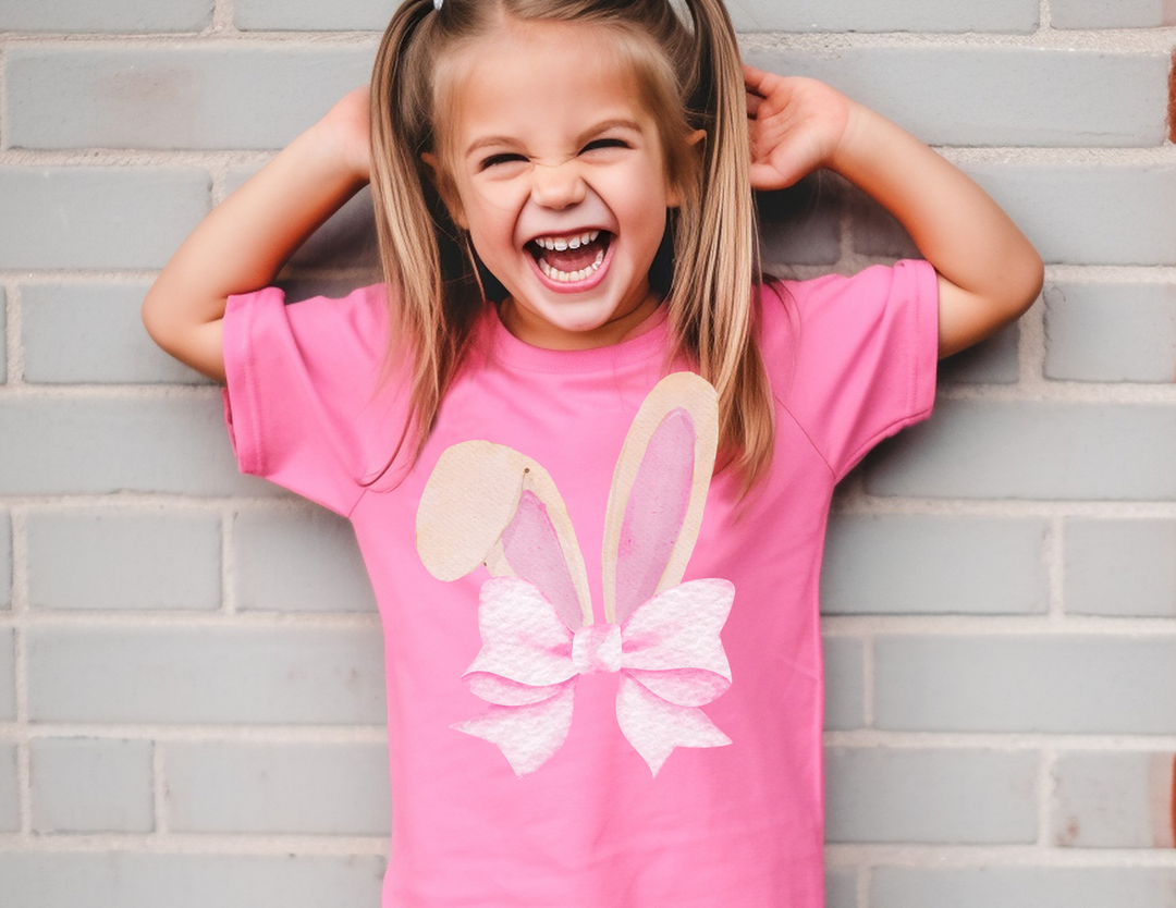 Young girl with pigtails joyfully poses, wearing Bunny Bow Toddler Tee, featuring a stylized bunny-ear graphic, highlighting its softness and perfect fit for toddlers.