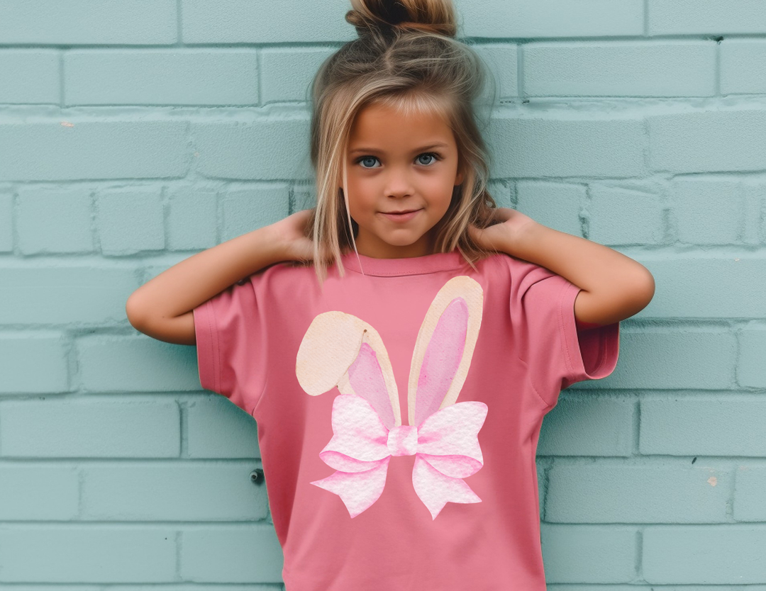 Young girl in a pink Bunny Bow Kids Tee stands against a pale teal brick wall, showcasing the shirt’s watercolor bunny ears and bow graphic.