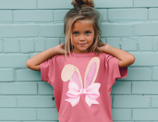 Young girl in a pink Bunny Bow Kids Tee stands against a pale teal brick wall, showcasing the shirt’s watercolor bunny ears and bow graphic.