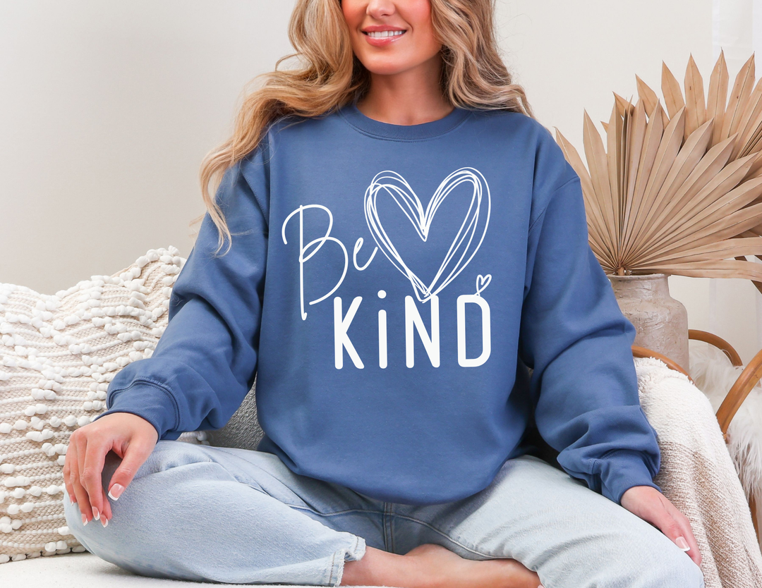 Person in slate-blue Be Kind Crew sweatshirt with heart graphic, seated on couch with light-washed jeans, textured pillow, and natural décor nearby.