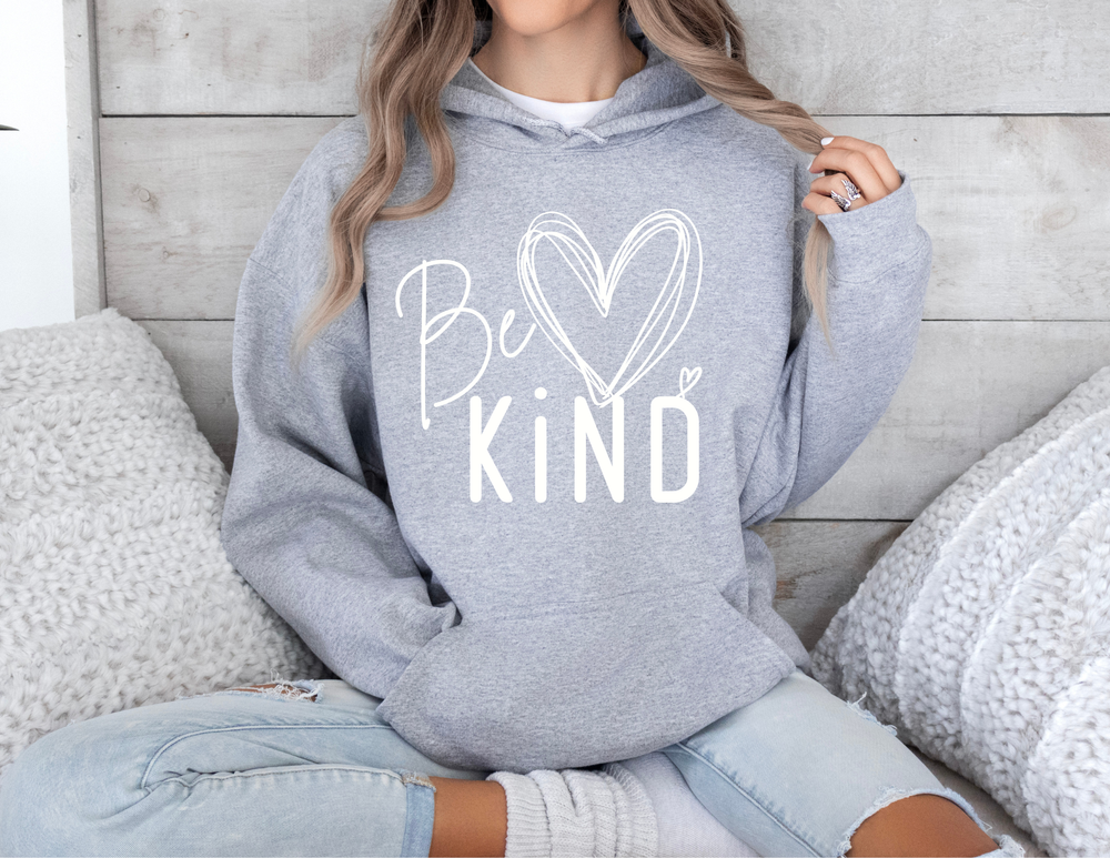 Person wearing a Be ♥ KiND heather-gray youth hoodie, with light-wash ripped jeans, sitting cross-legged, surrounded by chunky knit cushions. Face not visible.