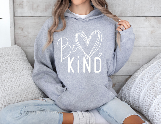 Person wearing a Be ♥ KiND heather-gray youth hoodie, with light-wash ripped jeans, sitting cross-legged, surrounded by chunky knit cushions. Face not visible.