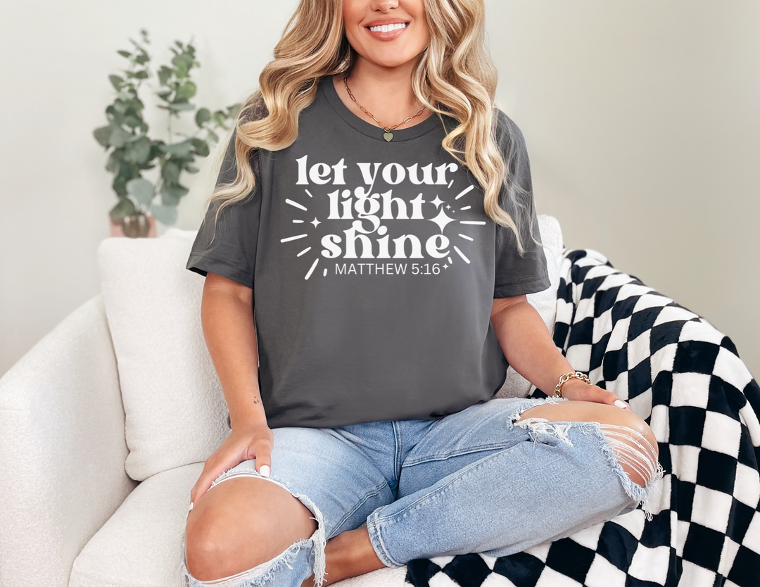 Person wearing Let Your Light Shine tee in dark gray, paired with ripped jeans, sitting on a sofa. The T-shirt displays a sparkle design and biblical reference.