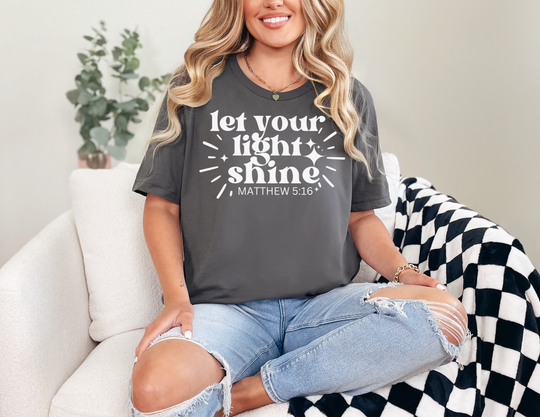 Person wearing Let Your Light Shine tee in dark gray, paired with ripped jeans, sitting on a sofa. The T-shirt displays a sparkle design and biblical reference.