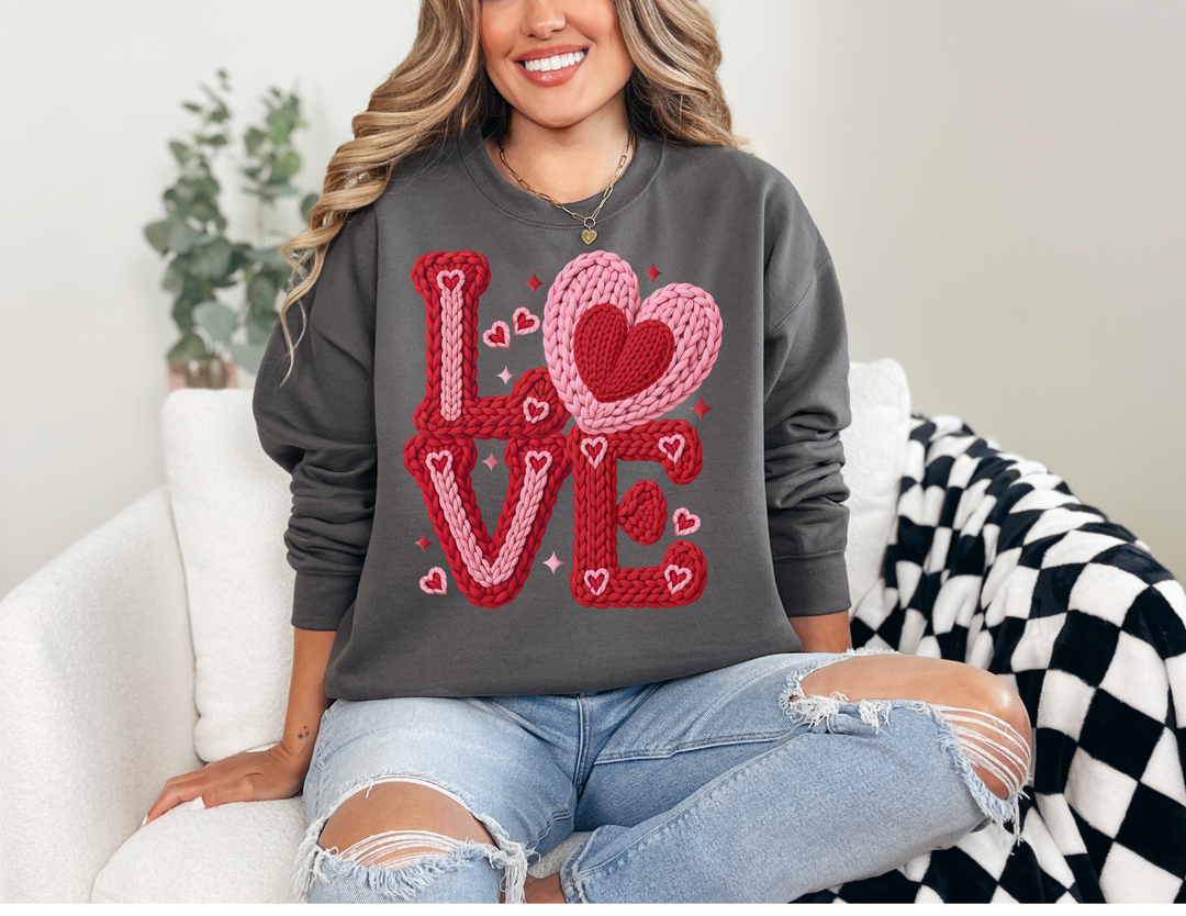 Woman in ripped jeans wearing a charcoal Love Heart Long Sleeve Tee with red and pink knitted text, seated in a cream armchair.