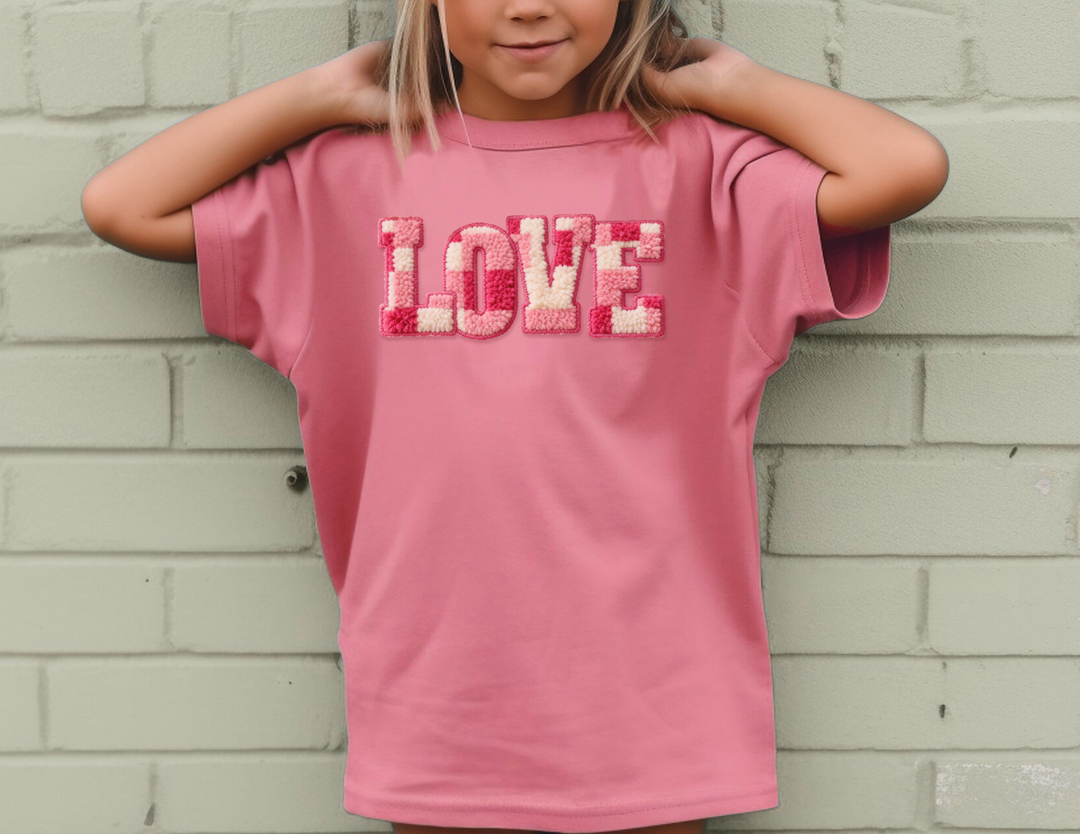 Child wearing a pink LOVE Kids Tee with tufted letters, standing against a brick wall, arms bent behind head, showcasing a comfortable, durable design.