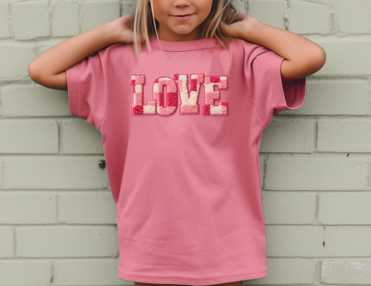 Child wearing a pink LOVE Kids Tee with tufted letters, standing against a brick wall, arms bent behind head, showcasing a comfortable, durable design.