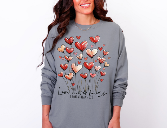 Person wearing the Love Never Fails long sleeve tee, featuring heart-shaped flowers and Bible reference, offering comfort and style in 100% ring-spun cotton.