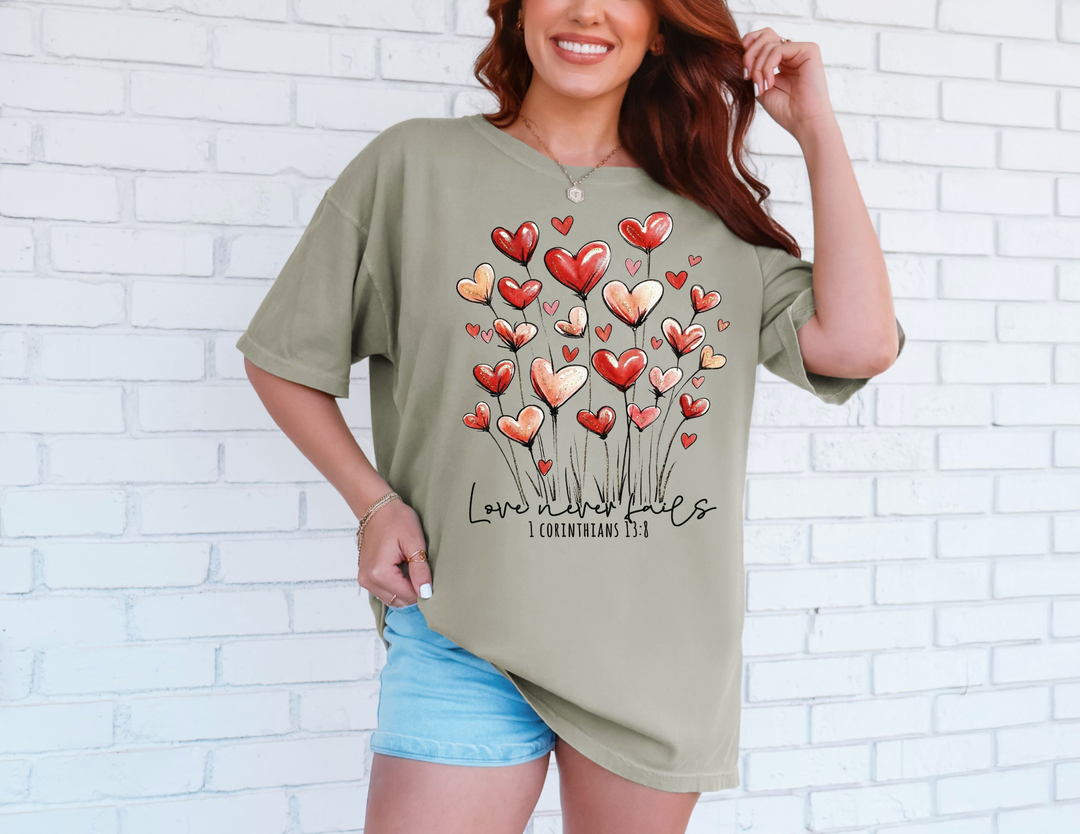 Woman in sage-green Love Never Fails tee with heart-shaped flowers, paired with light denim shorts, against a white brick wall.