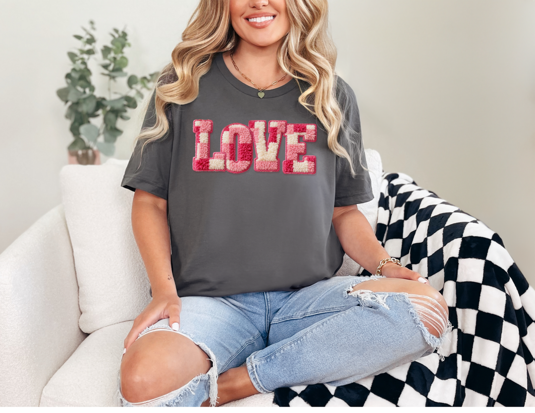 Person wearing Love Tee in dark gray, featuring pink, cream, and red tufted lettering, paired with light-wash jeans and simple jewelry, seated on a chair.