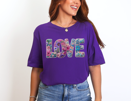 Person wearing vibrant “LOVE” Spring Tee with colorful floral design, paired with denim jeans, necklace, and bracelet, embodying casual, cheerful style.