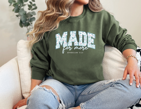 Person wearing a forest-green Made for More Crew sweatshirt with leaf-and-berry pattern, paired with light-wash jeans, showcasing simple jewelry.