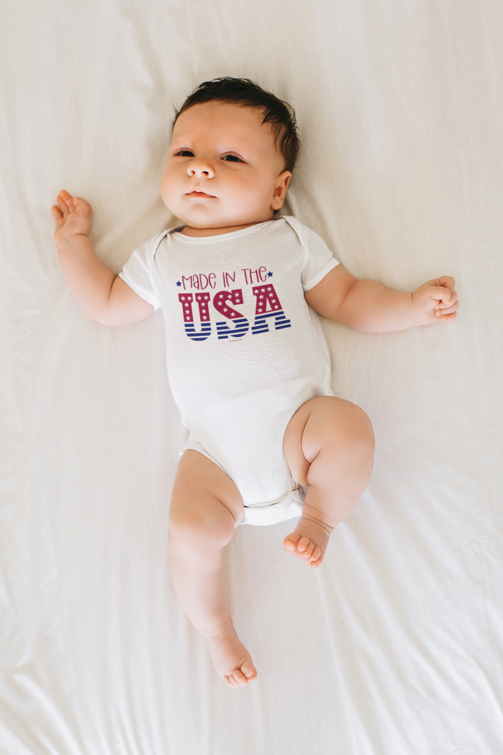 A baby lies on a bed, wearing a Made in the USA Onesie from Worlds Worst Tees. This infant fine jersey bodysuit is 100% cotton, with ribbed knitting for durability and plastic snaps for easy changing access.