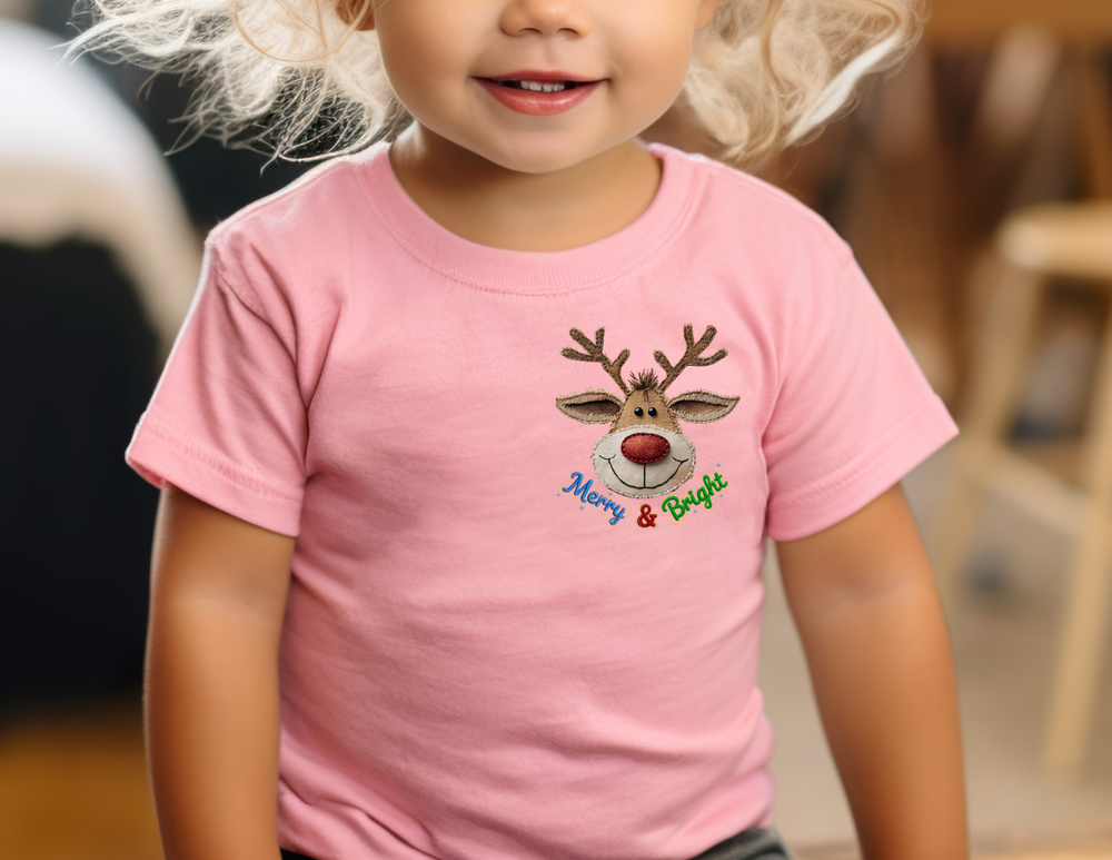 Child wearing Merry and Bright Kids Tee with a smiling reindeer graphic and embroidered text, showcasing a light pink, durable cotton T-shirt from Faith Family Designs.