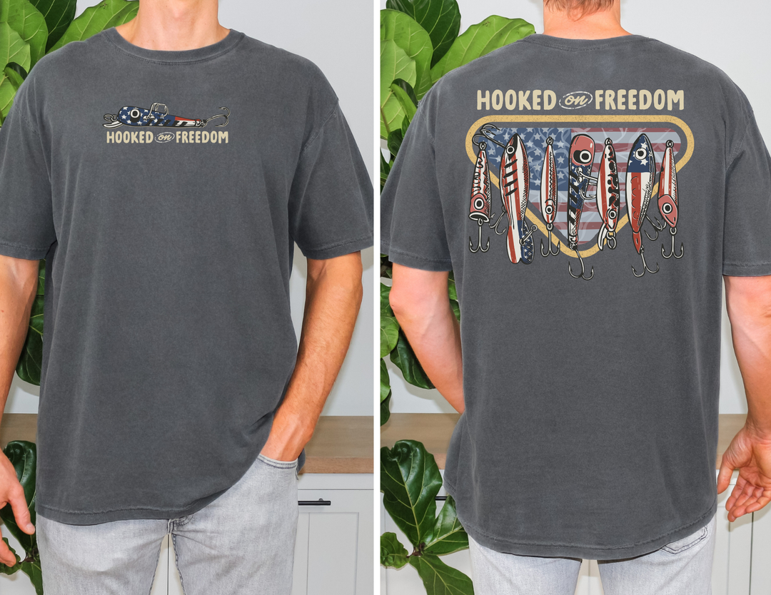Hooked on Freedom Tee featuring patriotic fishing lures and HOOKED on FREEDOM text on vintage-washed dark-gray, short-sleeve T-shirt, modeled indoors.