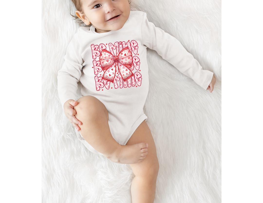 Baby in a soft Be Mine Onesie, featuring a pink-and-red bow design with text pattern, lying on a fluffy surface, smiling gently.
