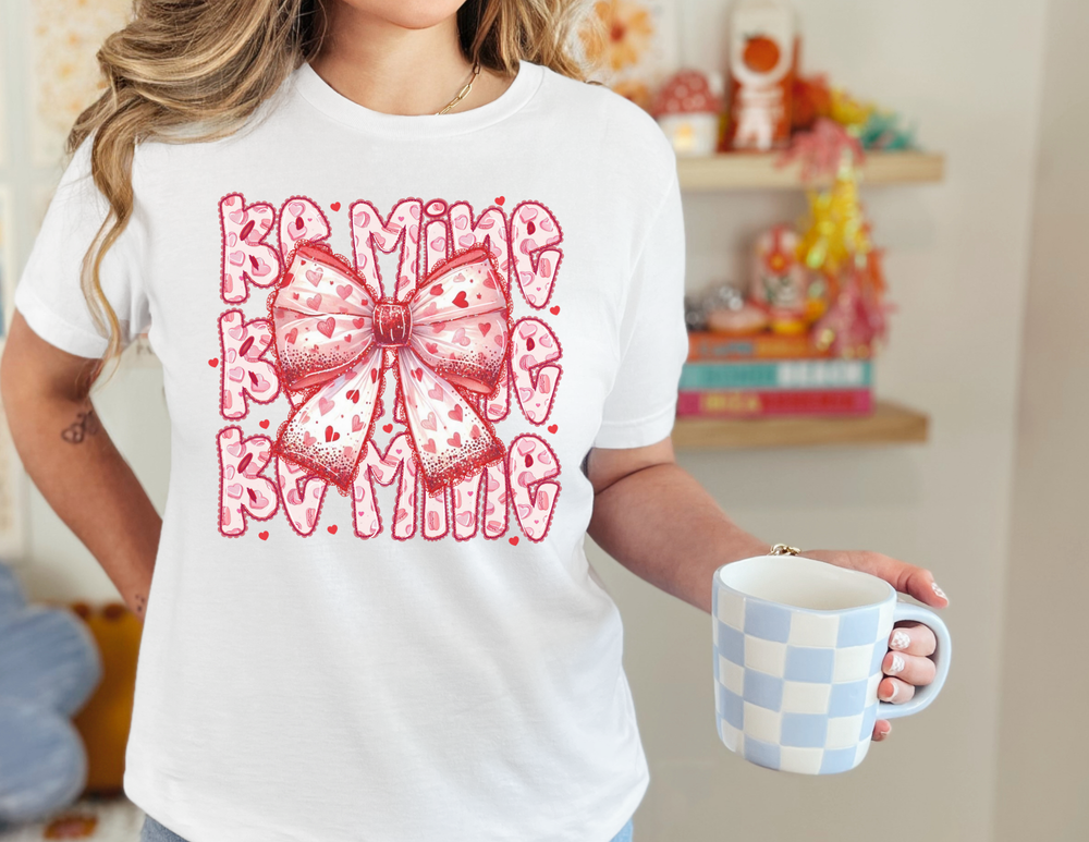 Person wearing Be Mine Tee featuring a pink-and-white bow with red hearts, holding a checkerboard mug. Casual, soft jersey perfect for everyday wear.