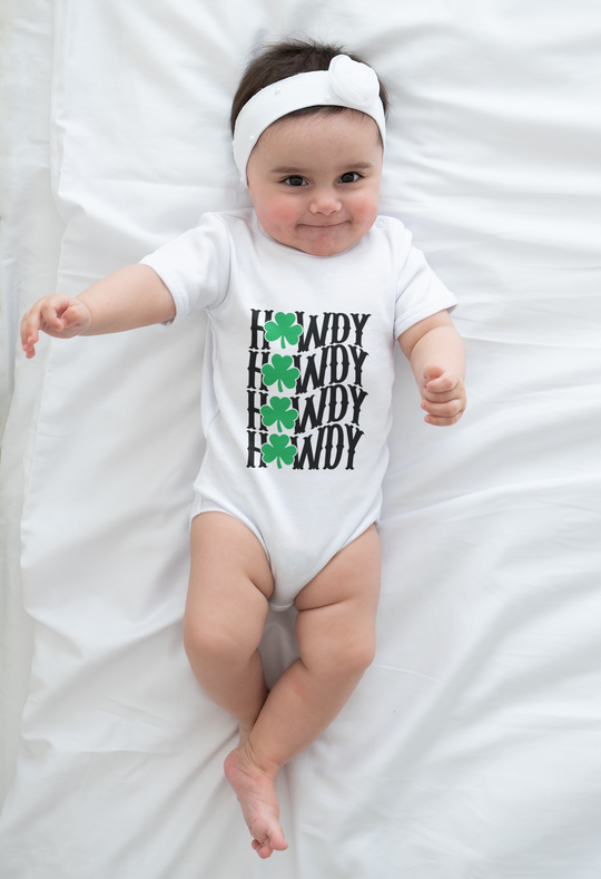 Baby wearing the Howdy Lucky Onesie, made from soft ring-spun cotton, lying on a white sheet with visible lap shoulders for easy adjustments.
