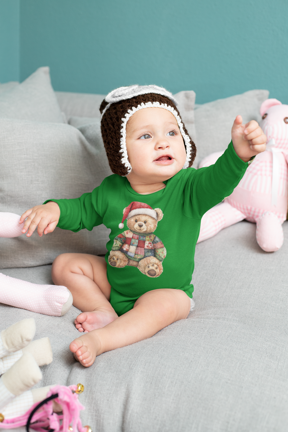 Baby wearing Christmas Bear Onesie with Santa hat graphic, sitting on a couch surrounded by soft toys, reaching out with one arm.