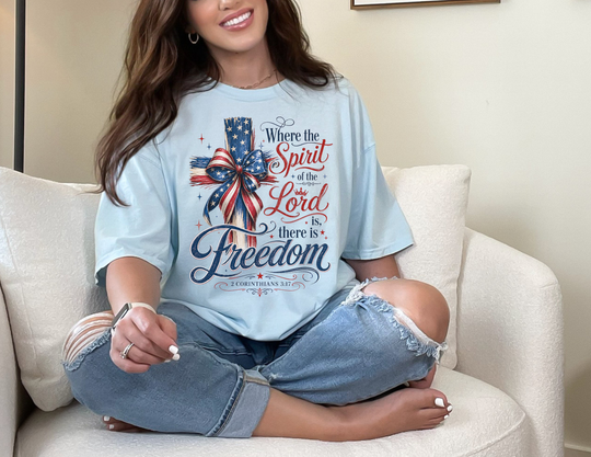 Young woman in a Spirit of the Lord Tee with a cross and bow graphic, sitting cross-legged on a sofa, wearing ripped jeans and holding a smartphone.