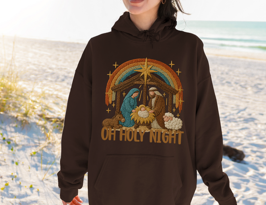 Person on beach wearing Oh Holy Night hoodie featuring embroidered nativity scene with Mary, Joseph, baby Jesus, star, donkey, sheep, and rainbow arch. Cozy, unisex design.