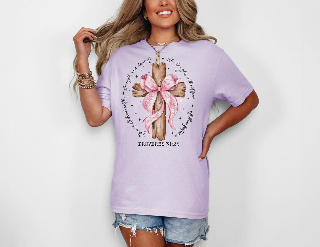 Woman in a light-lavender Proverbs 31:25 Tee featuring a wooden cross with a pink bow and biblical quote, paired with denim cutoff shorts.
