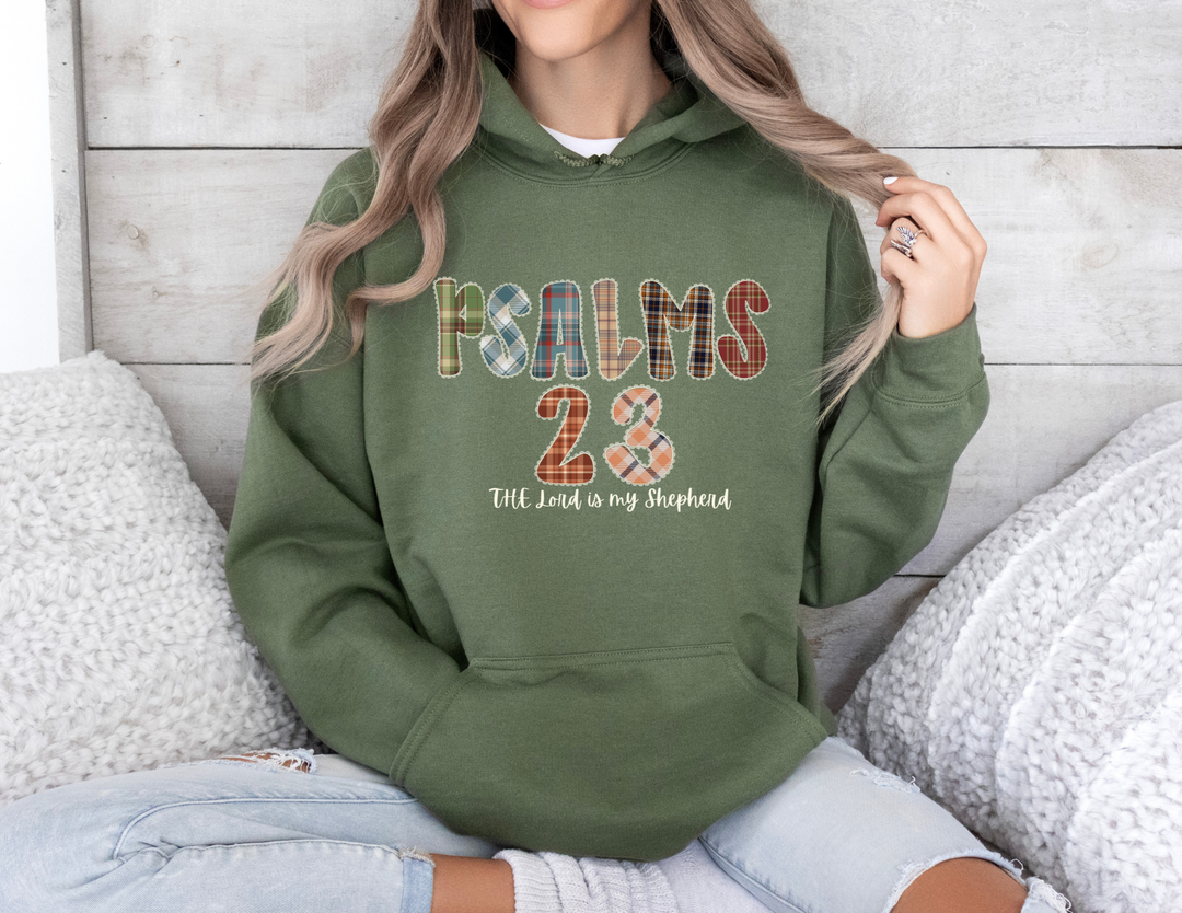 Person seated, wearing a forest-green Psalms 23 Hoodie with plaid lettering, paired with light-wash jeans. Cozy setting with knitted pillows, reflecting casual comfort.