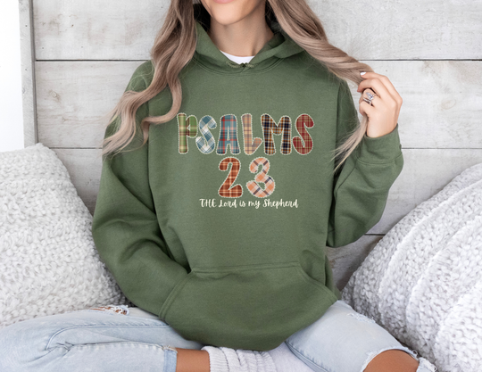 Person seated, wearing a forest-green Psalms 23 Hoodie with plaid lettering, paired with light-wash jeans. Cozy setting with knitted pillows, reflecting casual comfort.