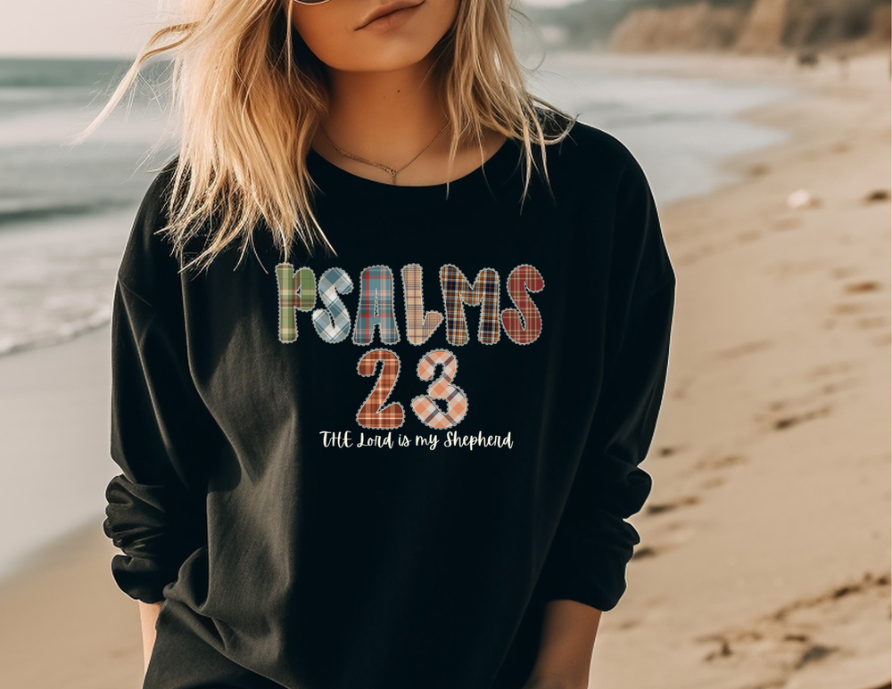 Person on beach wearing PSALMS 23 black sweatshirt with script The Lord is my Shepherd, showcasing Faith Family Designs' cozy, garment-dyed, 100% cotton long-sleeve tee.