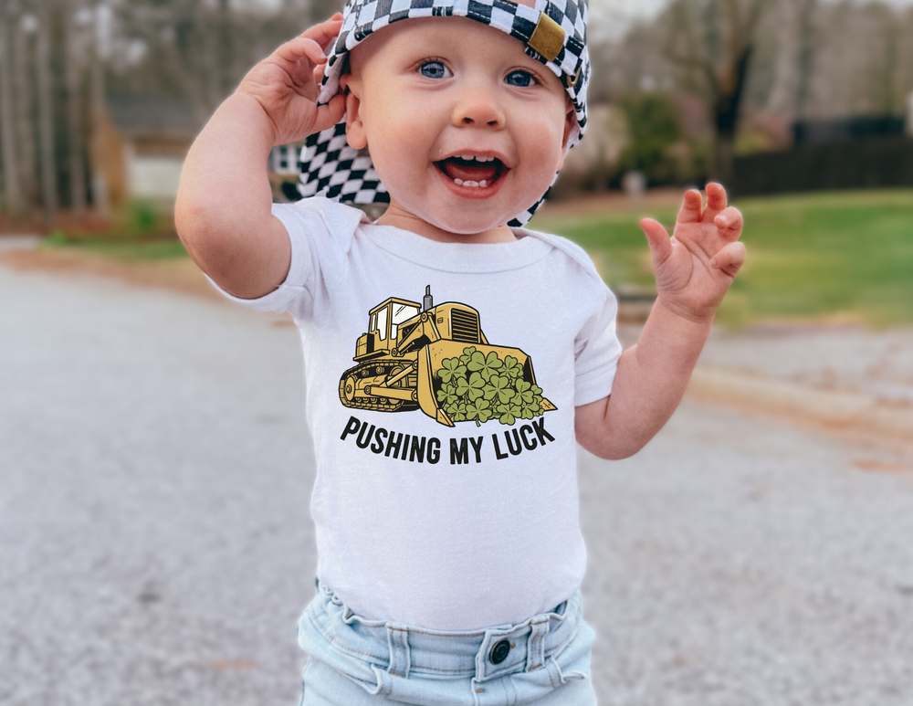 Toddler in a Pushing My Luck baby tee with bulldozer graphic, wearing a checkered cap, smiling outdoors. Ideal for comfort and durability from Faith Family Designs.