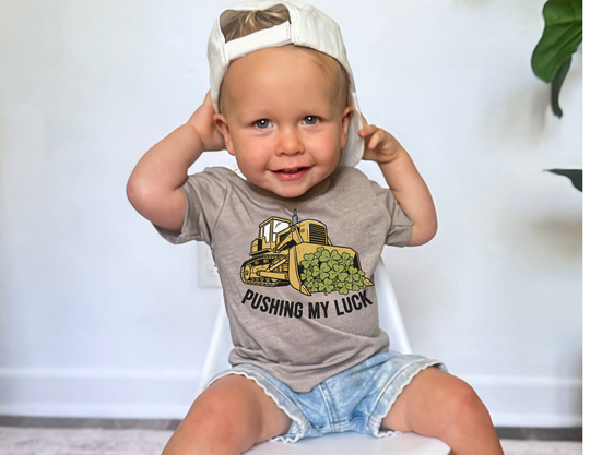 Toddler wearing Pushing My Luck Tee with bulldozer graphic and text, seated on a chair, showcasing comfort and playful design from Faith Family Designs.