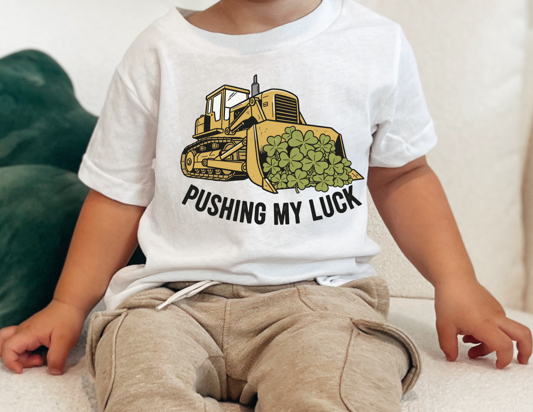 Child wearing Pushing My Luck Kids Tee with a cartoon bulldozer design, seated on a couch, showcasing the shirt's comfortable fit and playful graphic.