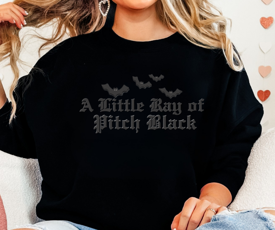 Person wearing black sweatshirt with four small bats and A Little Ray of Pitch Black in gothic font, embodying unique graphic style from Faith Family Designs.