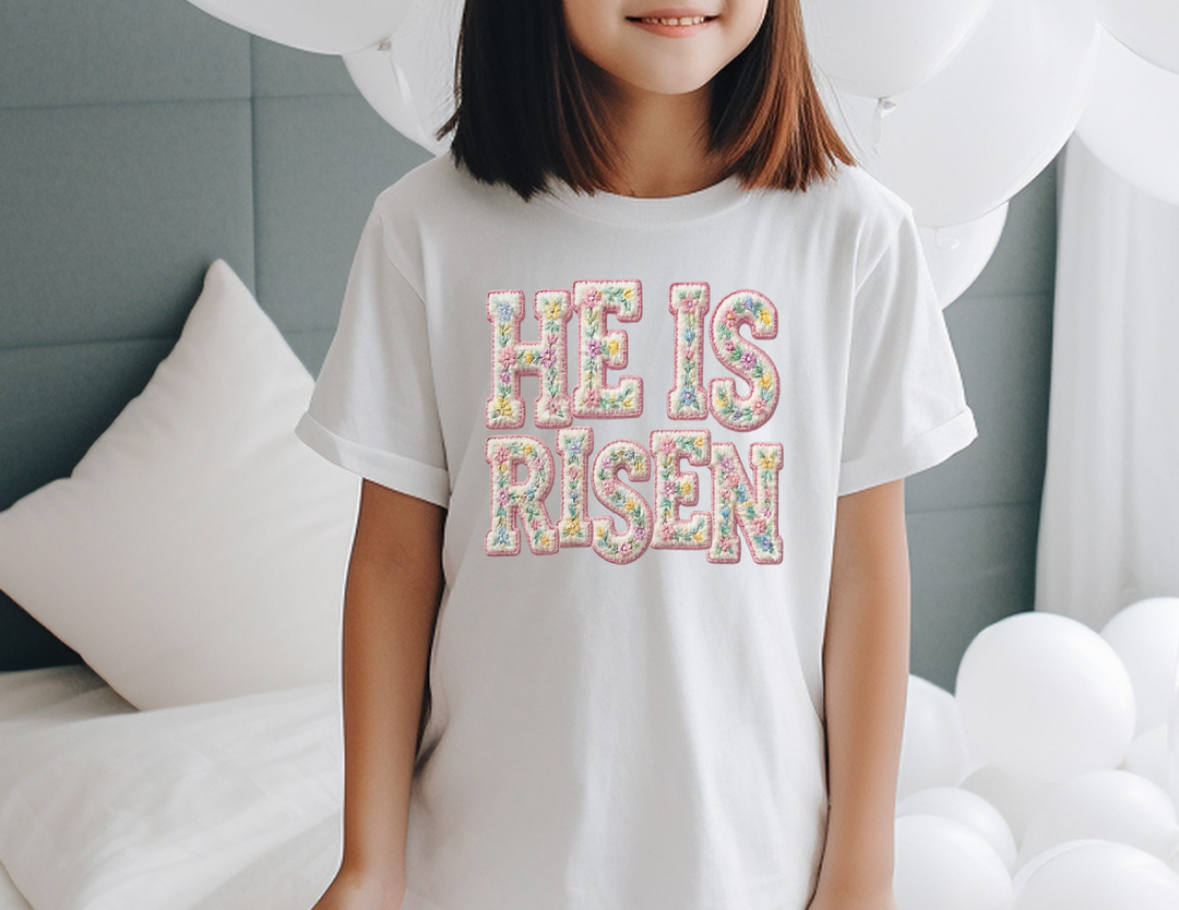 Child wearing HE IS RISEN white tee with pastel floral letters, embodying comfort and agility. Designed by Faith Family Designs for active kids' lifestyles.