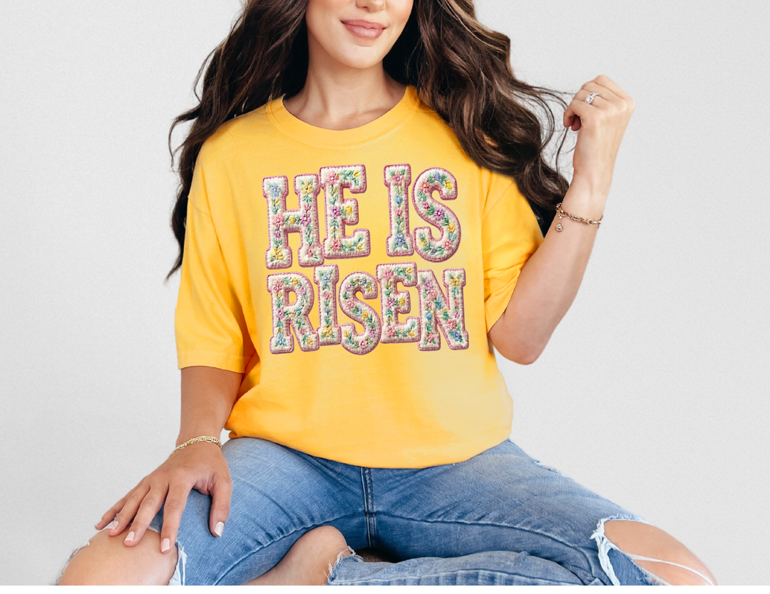 A person wears the He is Risen tee, featuring floral letters on a yellow background, paired with ripped blue jeans.