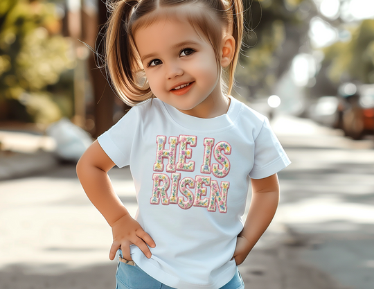 Toddler wearing He is Risen tee with floral letters, smiling outdoors. Ideal for sensitive skin, classic fit, perfect for early adventures.