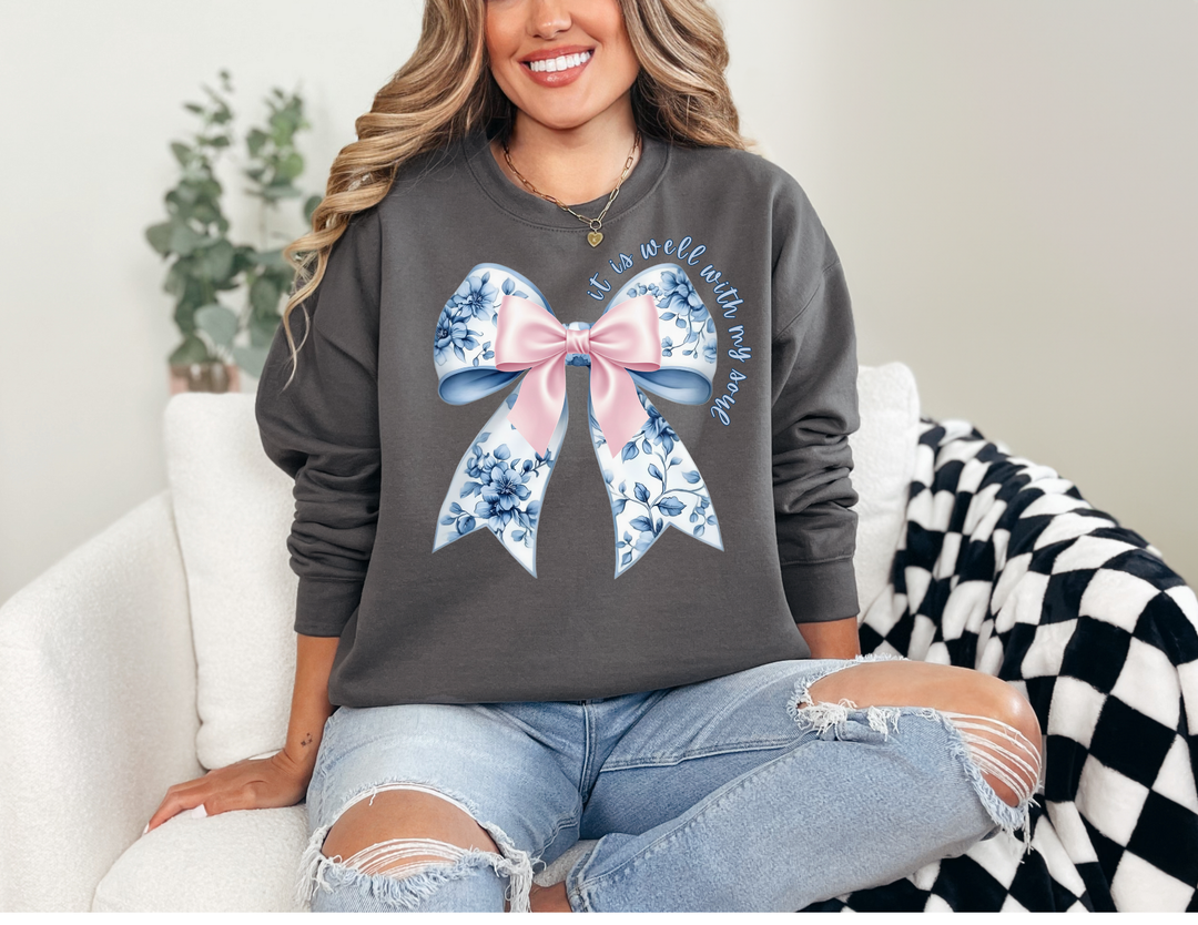 Woman wearing It is Well With My Soul Crew sweatshirt, featuring a floral bow design, sitting casually on a sofa with light-wash, ripped jeans.