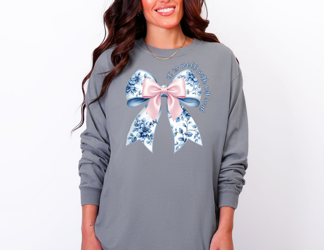 Person wearing a loose-fit slate-gray long-sleeve shirt with a stylized bow and the phrase “It is well with my soul.”