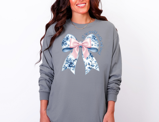 Person wearing a loose-fit slate-gray long-sleeve shirt with a stylized bow and the phrase “It is well with my soul.”