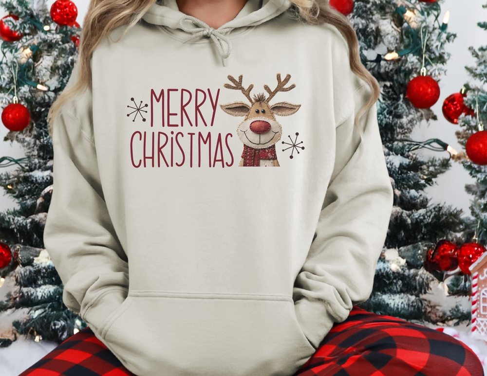 Person wearing a MERRY CHRISTMAS Reindeer Hoodie and buffalo-plaid pajama bottoms, sitting by a decorated Christmas tree with a gingerbread-style house ornament.