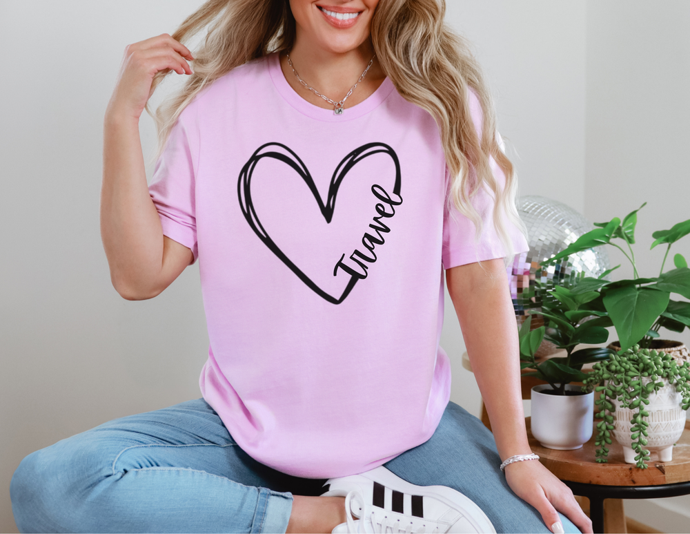 Woman in light-blue jeans wearing a pink Travel Tee with heart design, sitting cross-legged in a cozy interior with potted plants and a disco ball.