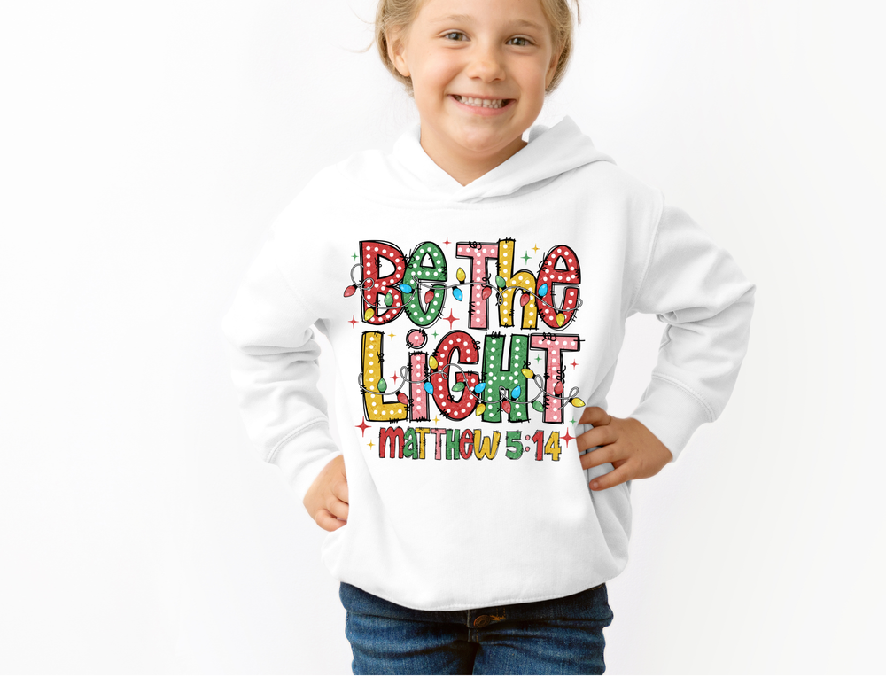 Child smiling in a Be the Light Youth Hoodie with colorful hand-lettered design and kangaroo pocket, embodying Faith Family Designs' unique and comfortable style.