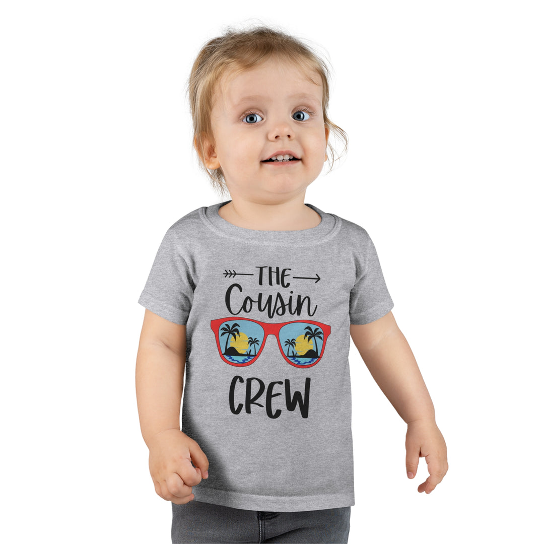 A toddler wearing a Cousin Crew Toddler Tee, featuring a grey shirt with black letters. Classic fit, 100% Ringspun cotton, durable double-needle collar, sleeve, and bottom hems. Ideal for personalized designs.