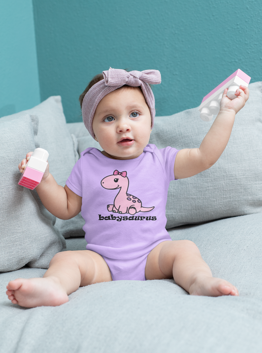 A baby girl wearing a purple onesie with a cartoon dinosaur design, held by a baby's hand. Infant fine jersey bodysuit made of 100% cotton, featuring ribbed bindings and plastic snaps for easy changing.