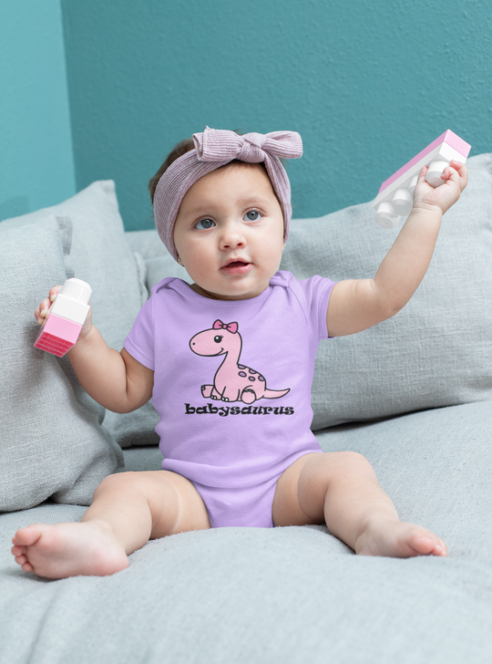 A baby girl wearing a purple onesie with a cartoon dinosaur design, held by a baby's hand. Infant fine jersey bodysuit made of 100% cotton, featuring ribbed bindings and plastic snaps for easy changing.