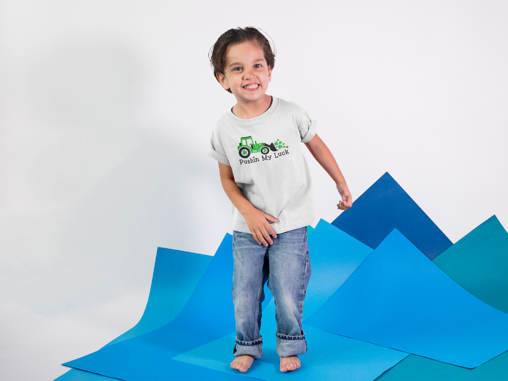A child in a white shirt and jeans stands on a blue mat, embodying playful innocence in the Pushin My Luck Kids Tee. Made of 100% combed ringspun cotton, this tee offers a soft, high-quality feel.