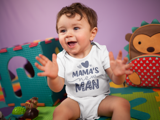 A baby boy sits happily surrounded by toys, wearing a Mama's New Man Onesie from Worlds Worst Tees. This infant bodysuit is 100% cotton, featuring ribbed knitting for durability and plastic snaps for easy changing access.