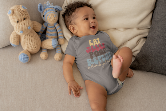 A baby lies on a couch surrounded by stuffed animals, wearing the Eat Sleep Poop Onesie from Worlds Worst Tees. This 100% cotton bodysuit features ribbed bindings and plastic snaps for easy changes.