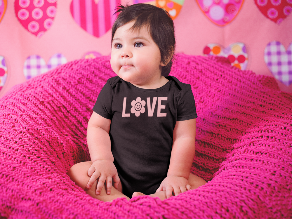 A baby in a pink blanket sits comfortably, showcasing the Love Onesie from Worlds Worst Tees. This infant fine jersey bodysuit is 100% cotton, featuring ribbed knitting for durability and easy plastic snaps for changing.
